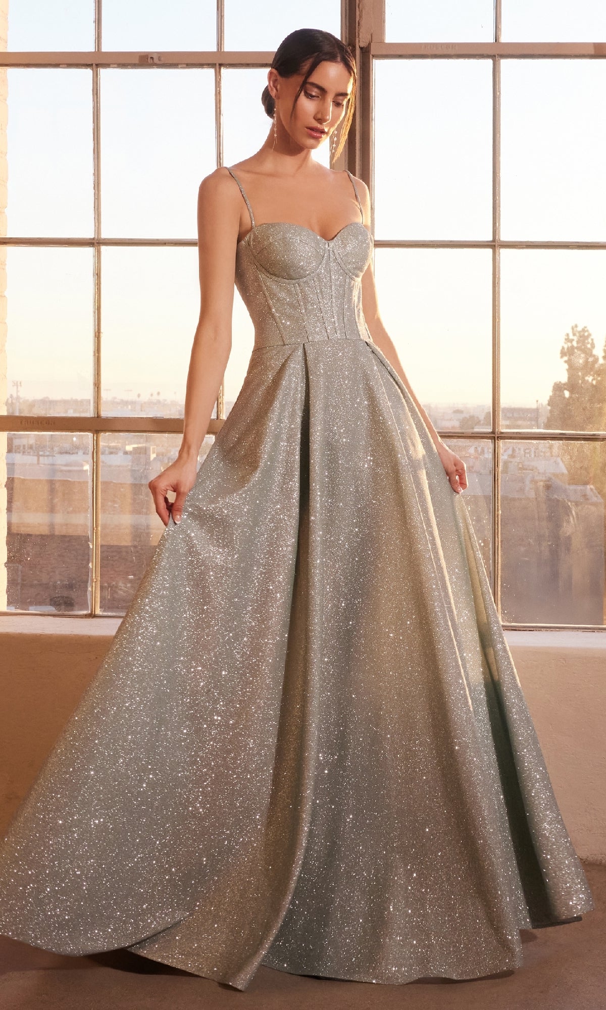 A woman stands indoors by large windows, wearing the Amoris T706 Lace-Up Long Glitter Prom Ball Gown in light green, featuring a fitted bodice, thin straps, and a sparkling floor-length skirt.